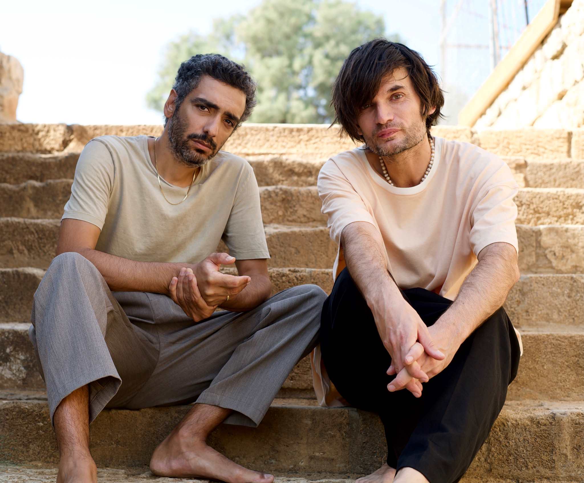 Dudu Tassa (left) and Jonny Greenwood sitting on stone steps, the portrait issued for Jarak Qaribak, 2023