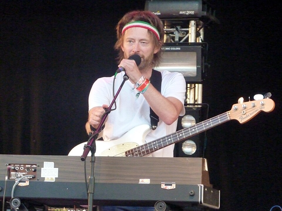Thom Yorke in a red, white and green sweatband at a microphone, a white bass slung over a keyboard, on stage at Glastonbury, 25 June 2010